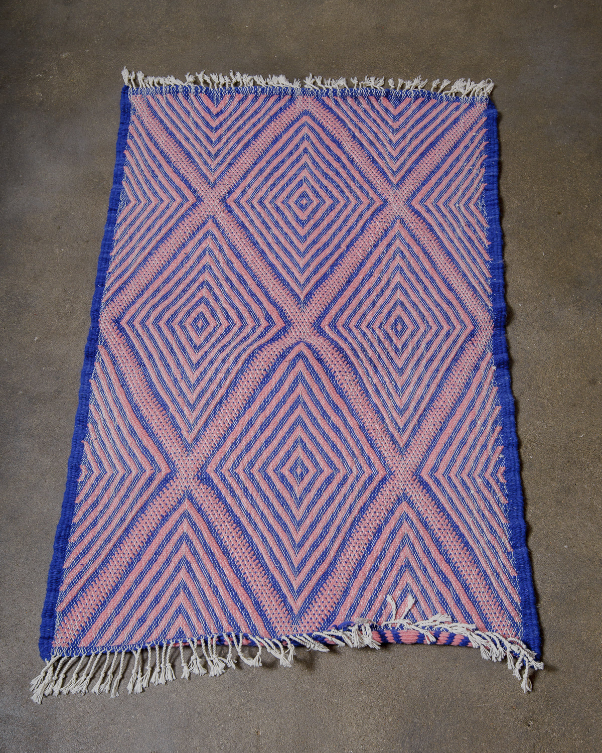 Pink and blue Kelim rug with some curling at the ends, laying on a concrete floor.